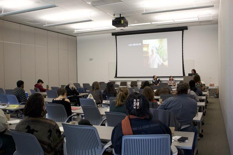 Photo of event attendees in a classroom looking at a presenter with a slide that says "Las Abuelas"