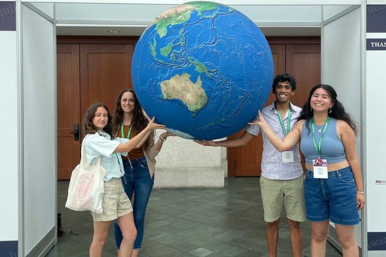 Photo of four people posing in front of a model of the earth with "Welcome to AAG 2024" above them