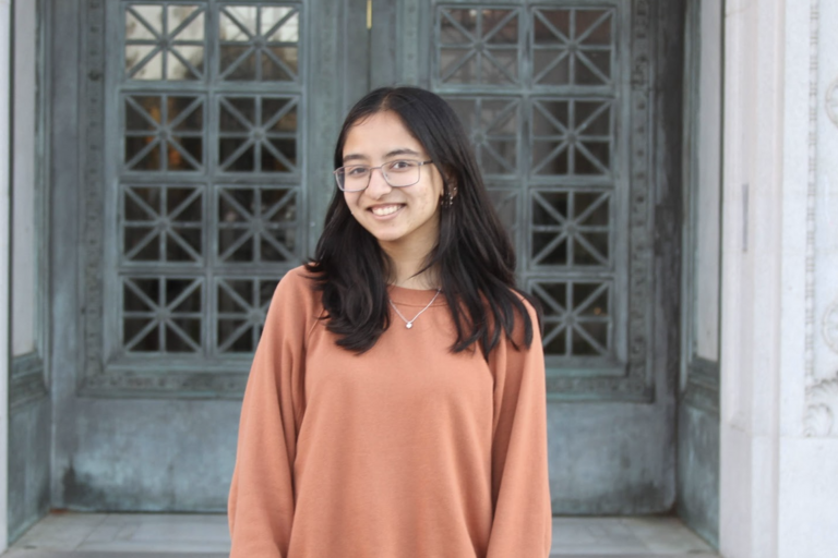 Young woman with black hair and glasses smiling in front of a blue patina door